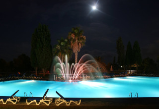 Illuminated swimming pool with fountains and palm trees at night at Camping Vilanova Park, Catalonia, Spain.