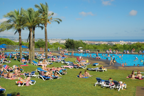 Personas descansando junto a la piscina en Camping Vilanova Park con vistas y palmeras en Cataluña, España.