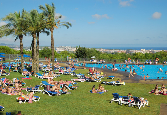 Personas descansando junto a la piscina en Camping Vilanova Park con vistas y palmeras en Cataluña, España.