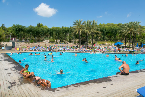 Gente disfrutando en la piscina al aire libre en Camping Vilanova Park, rodeada de palmeras, en Cataluña, España.