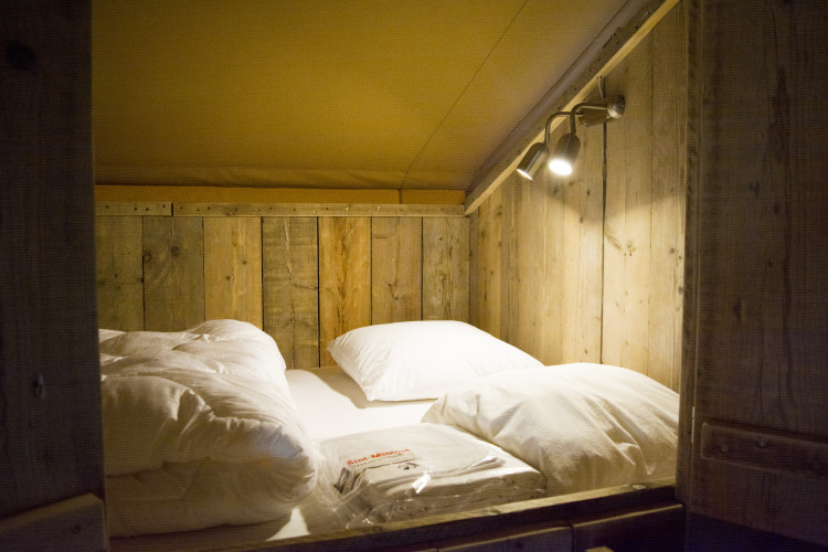 Cama acogedora con paredes de madera y ropa de cama dentro de una lujosa tienda de glamping en Bélgica.
