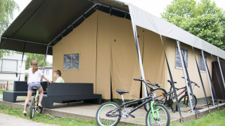 Niños juegan junto a una tienda safari glamping con bicicletas y muebles al aire libre en una terraza.