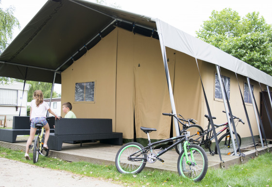 Niños juegan junto a una tienda safari glamping con bicicletas y muebles al aire libre en una terraza.