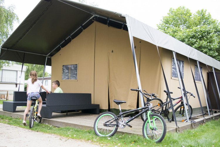 Niños juegan junto a una tienda safari glamping con bicicletas y muebles al aire libre en una terraza.