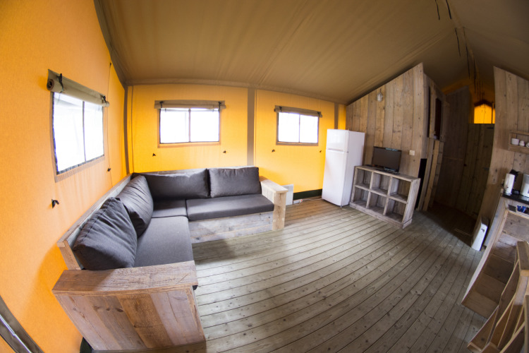 Vista interior de una tienda glamping safari con sofá, nevera y suelo de madera en Vakantiepark Klein Strand.