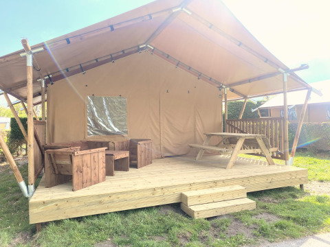 Safari-Zelt Glamping mit überdachter Holzterrasse, Holzmöbeln und Picknicktisch bei Sonnenschein.