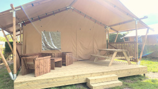 Safari-Zelt Glamping mit überdachter Holzterrasse, Holzmöbeln und Picknicktisch bei Sonnenschein.