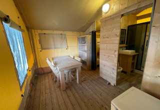 Interior de una tienda safari con muebles de madera, mesa de comedor, cocina y baño en Glamping Klein Strand.
