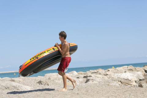 Un ragazzo porta un gommone sulla spiaggia rocciosa al Camping Vilanova Park, Catalogna, Spagna.