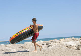 Un ragazzo porta un gommone sulla spiaggia rocciosa al Camping Vilanova Park, Catalogna, Spagna.