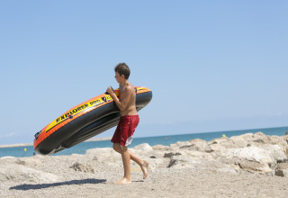 A boy carries an inflatable boat on a rocky beach with sea view at Camping Vilanova Park, Catalonia, Spain.