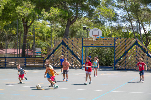 Niños jugando fútbol en una cancha de baloncesto al aire libre en Camping Vilanova Park, entre árboles verdes.