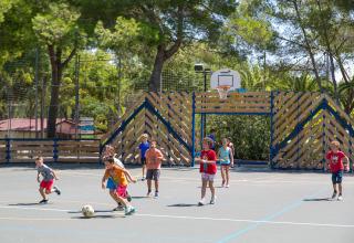 Children play football on an outdoor basketball court at Camping Vilanova Park, surrounded by lush trees.