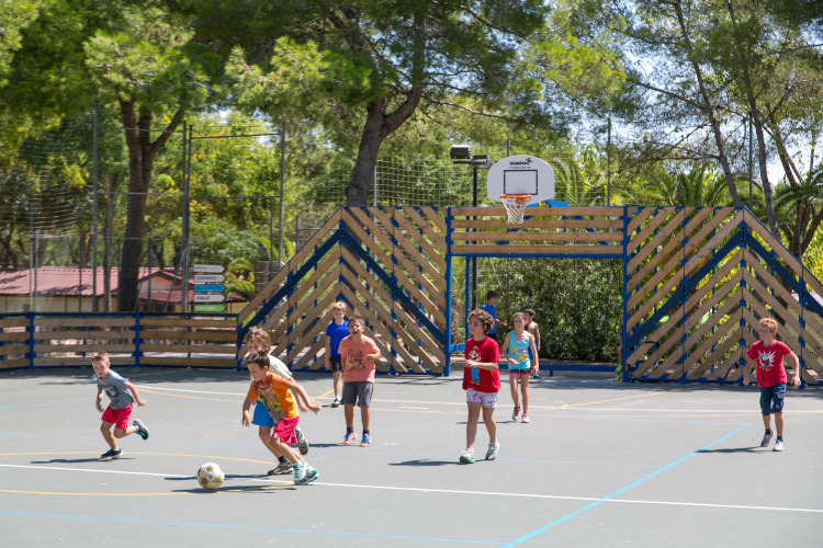 Kinderen spelen voetbal op een buitensportveld bij Camping Vilanova Park, omringd door bomen.