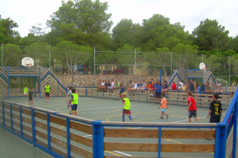 Niños juegan al balón en una cancha multideportiva con público en Camping Vilanova Park, Cataluña, España.
