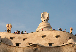 Tourists on the rooftop of a wavy stone building with sculptural chimneys under a clear blue sky.