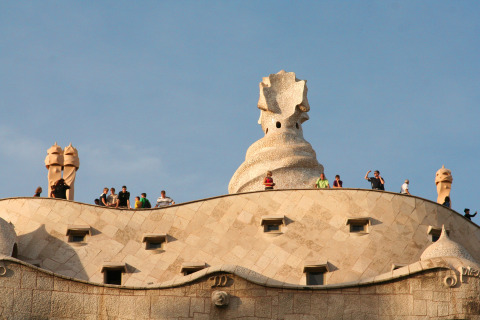 Tourists on the rooftop of a wavy stone building with sculptural chimneys under a clear blue sky.