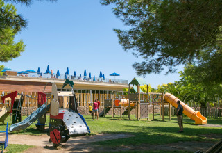 Sunny playground at Camping Vilanova Park in Spain with slides, play structures, trees, and blue umbrellas.