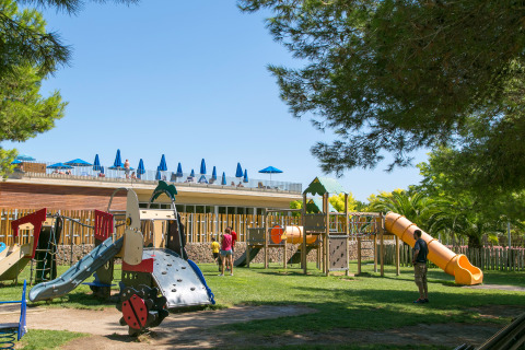 Kinderspeelplein in Camping Vilanova Park, met glijbanen, speeltoestellen en blauwe parasols op de achtergrond.