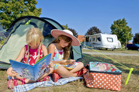 Twee meisjes lezen voor een tent op Camping Terme Catez, met caravan achter hen op een zonnige dag in Slovenië.