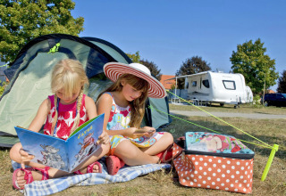 Dos niñas leyendo frente a una tienda en Camping Terme Catez, con caravana al fondo, día soleado en Eslovenia.