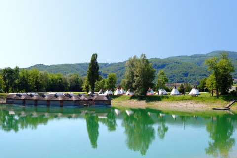 Lakeside view with wooden huts and tents at Camping Terme Catez, Slovenia, with hills in background.