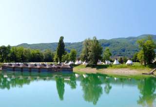 Lakeside view with wooden huts and tents at Camping Terme Catez, Slovenia, with hills in background.