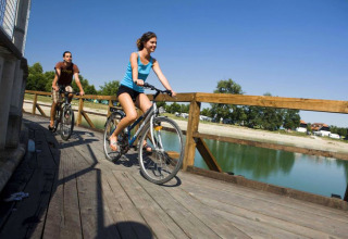 Two people cycle on a wooden bridge over water at Camping Terme Catez, Lower Sava, Slovenia, with blue skies.