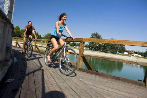 Two people cycle on a wooden bridge over water at Camping Terme Catez, Lower Sava, Slovenia, with blue skies.