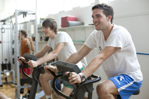 Twee jonge mannen fietsen op hometrainers in de fitnessruimte van Camping Terme Catez in Slovenië.