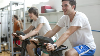 Dos hombres jóvenes usan bicicletas estáticas y sonríen en el gimnasio de Camping Terme Catez en Eslovenia.