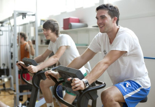 Deux jeunes hommes font du vélo d’appartement en souriant à la salle de sport du Camping Terme Catez en Slovénie.