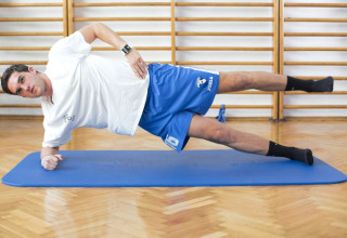 A man performs a side plank exercise on a blue mat in a gym with wooden floors and wall bars.