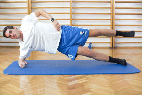 A man performs a side plank exercise on a blue mat in a gym with wooden floors and wall bars.