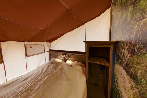 Vista interior de la tienda safari SunLodge Jungle con cama doble y estantería en Camping Vilanova Park, España.