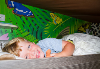 Un niño sonriente tumbado en la cama de una tienda safari con murales de mariposas en Vilanova Park, España.