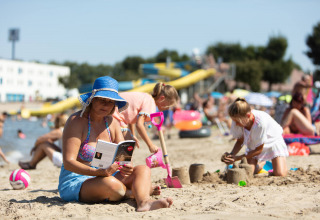 Woman reads a book on the beach at Vakantiepark Klein Strand, West Flanders, as kids play in sand.