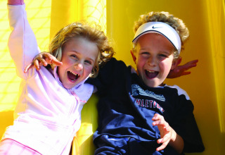 Two smiling children play together on a yellow slide at Vakantiepark Klein Strand in Belgium.