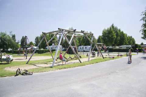 Kinder spielen auf einem Spielplatz mit Schaukeln und Klettergerüsten in einem Ferienpark mit Wohnwagen.