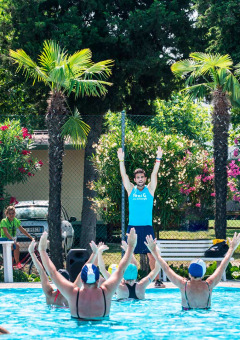 Instructor dirige una clase de gimnasia acuática en la piscina de Campeggio Gasparina, rodeada de palmeras y flores.