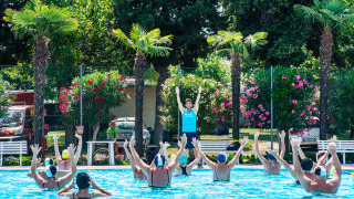 Instructor dirige una clase de gimnasia acuática en la piscina de Campeggio Gasparina, rodeada de palmeras y flores.