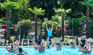 Instructor dirige una clase de gimnasia acuática en la piscina de Campeggio Gasparina, rodeada de palmeras y flores.