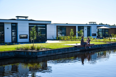 Due persone siedono vicino all’acqua davanti a case vacanza moderne a MarinaPark Residentie Nieuw Loosdrecht.