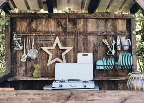 Cocina exterior con hornillo de gas, pared de madera, utensilios y vajilla azul en Camping d'Artagnan, Occitania.