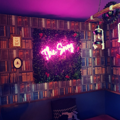 Rincón acogedor con papel tapiz de libros, letrero rosa neón 'The Snug', farol y plantas decorativas.