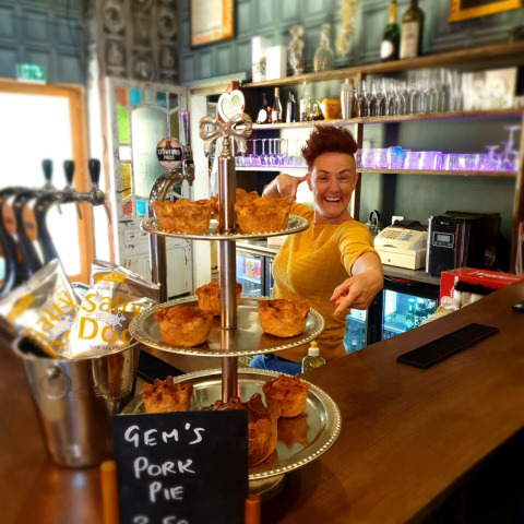 A cheerful person behind a bar points at trays of homemade pork pies with a sign reading 'Gem's Pork Pie'.