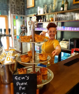 Une personne souriante derrière un bar désigne des plateaux de tourtes au porc avec une pancarte 'Gem's Pork Pie'.