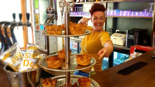 Une personne souriante derrière un bar désigne des plateaux de tourtes au porc avec une pancarte 'Gem's Pork Pie'.