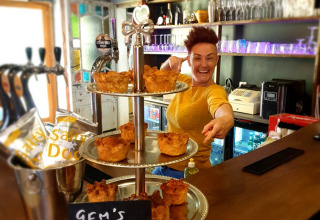 A cheerful person behind a bar points at trays of homemade pork pies with a sign reading 'Gem's Pork Pie'.
