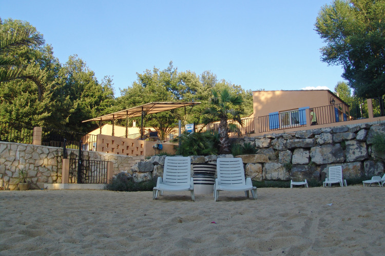 Sun loungers on a sandy area in front of a stone wall and holiday homes at Slow Village Provence Occitane, France.